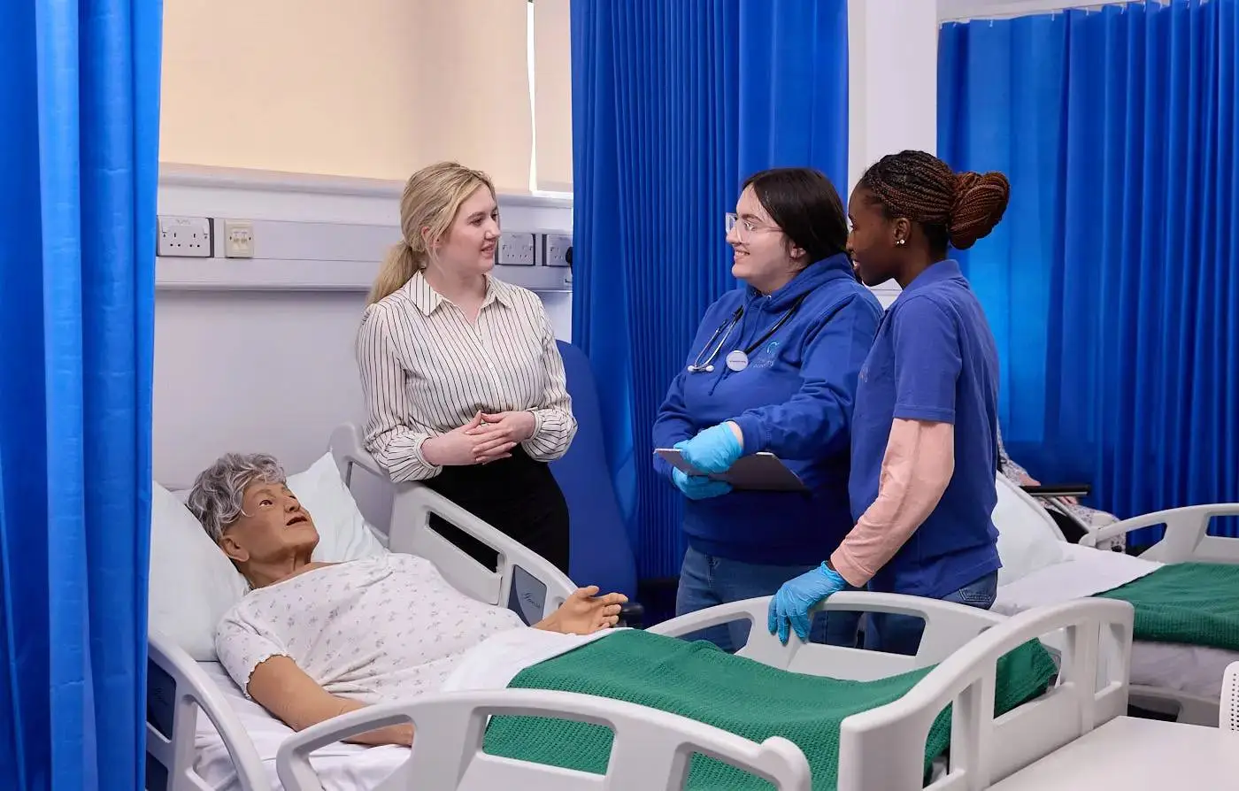 A woman in a hospital bed converses with three healthcare professionals, including one with a stethoscope, in a blue-curtained ward.