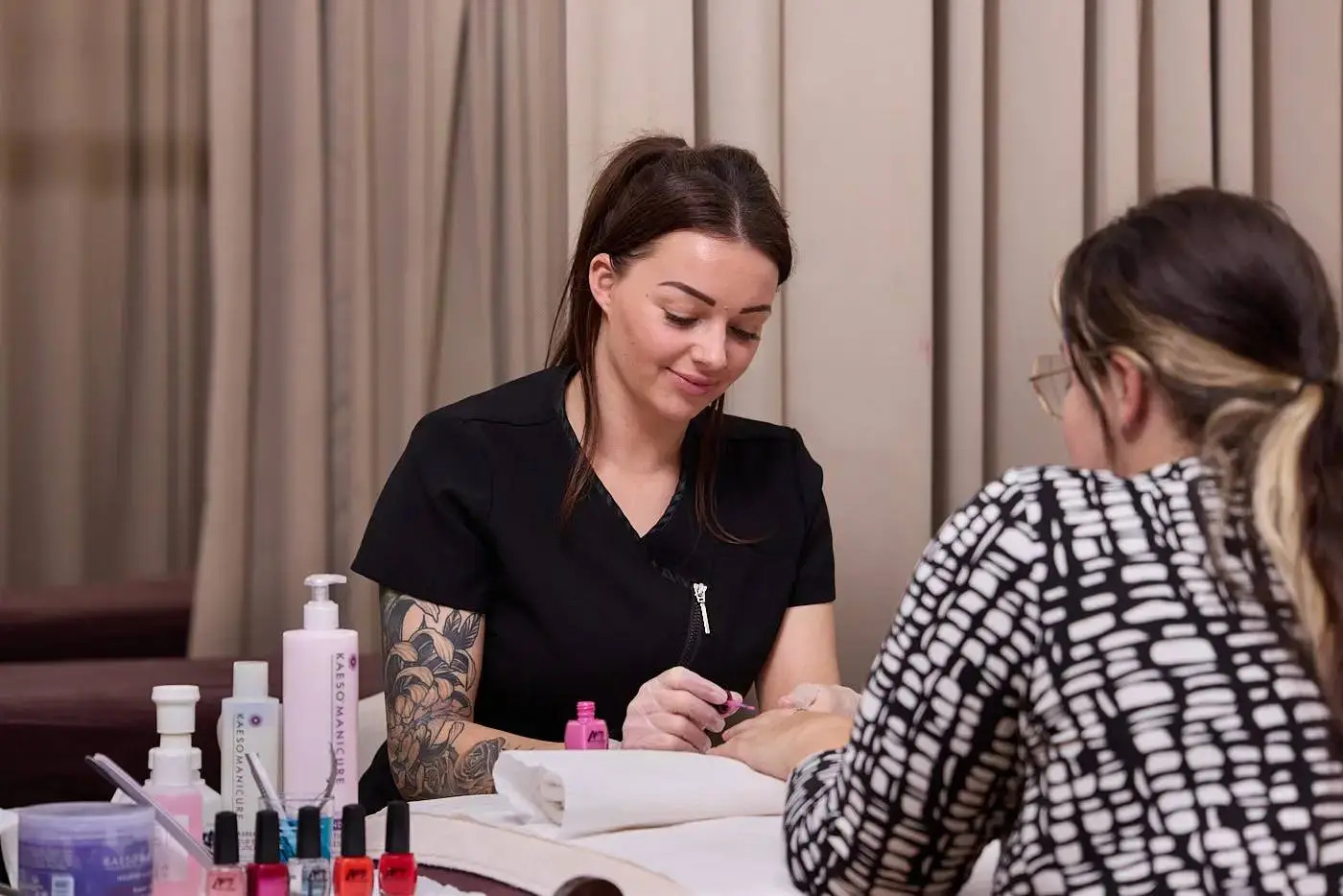 A woman in a black uniform paints another woman's nails in a nail salon setting.