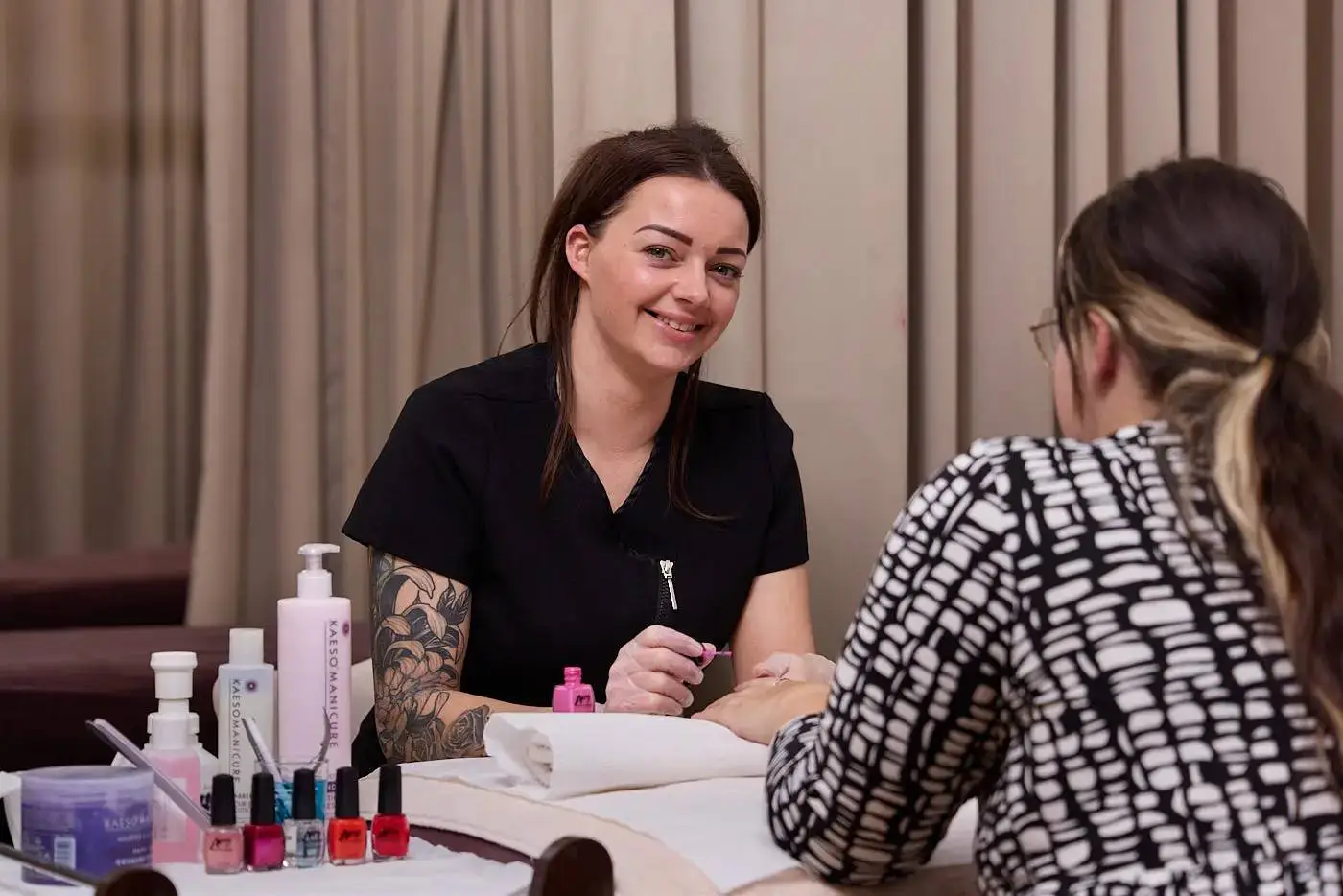A woman in a black uniform applying nail polish to another woman's hand in a nail salon setting.