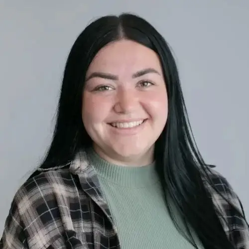 A smiling woman with long black hair wears a green top and a plaid shirt, standing against a plain background. She appears approachable and engaged, suitable for an educational context.