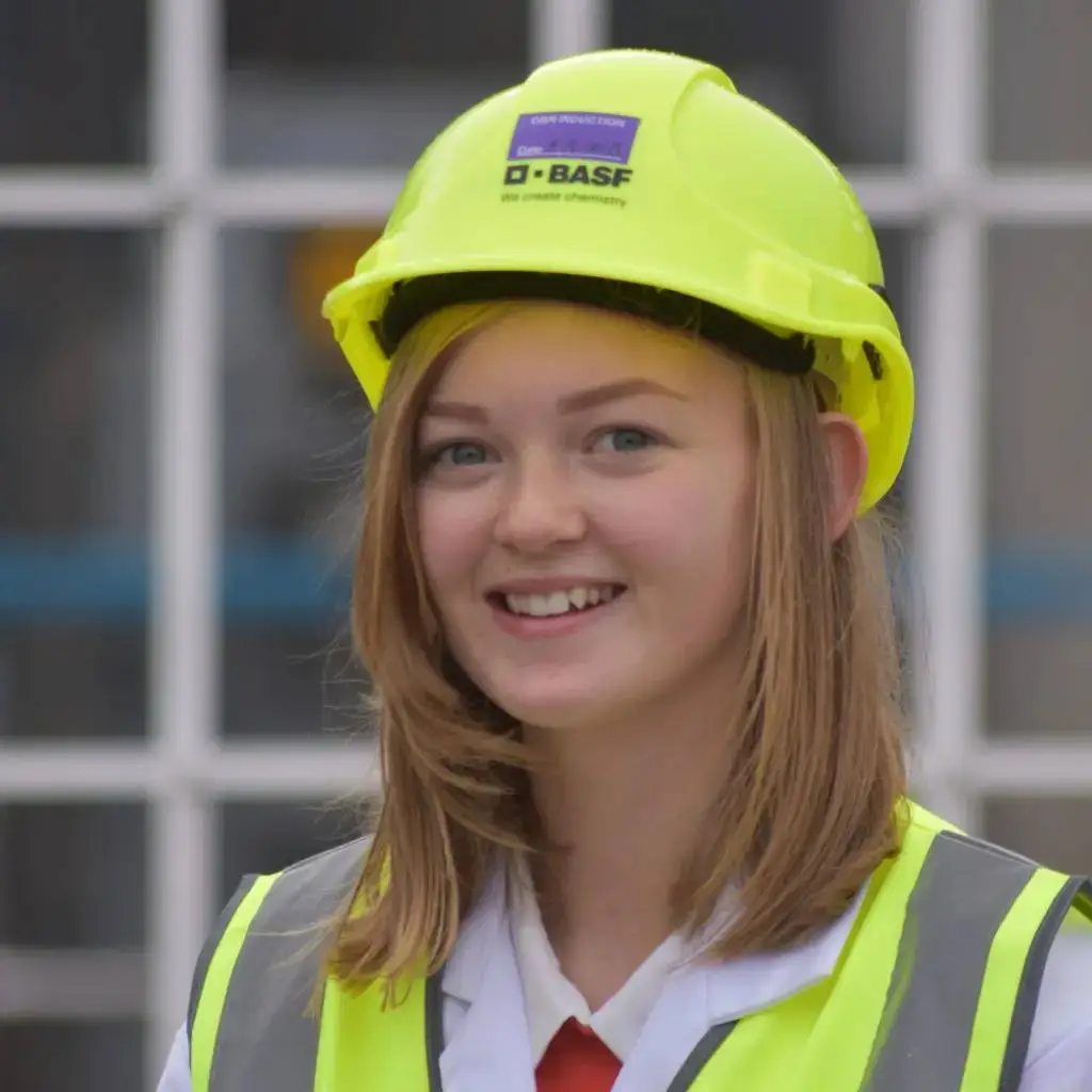 A young woman in a lab coat and high-visibility vest smiling, wearing a yellow hard hat with a BASF logo, standing in front of a window.