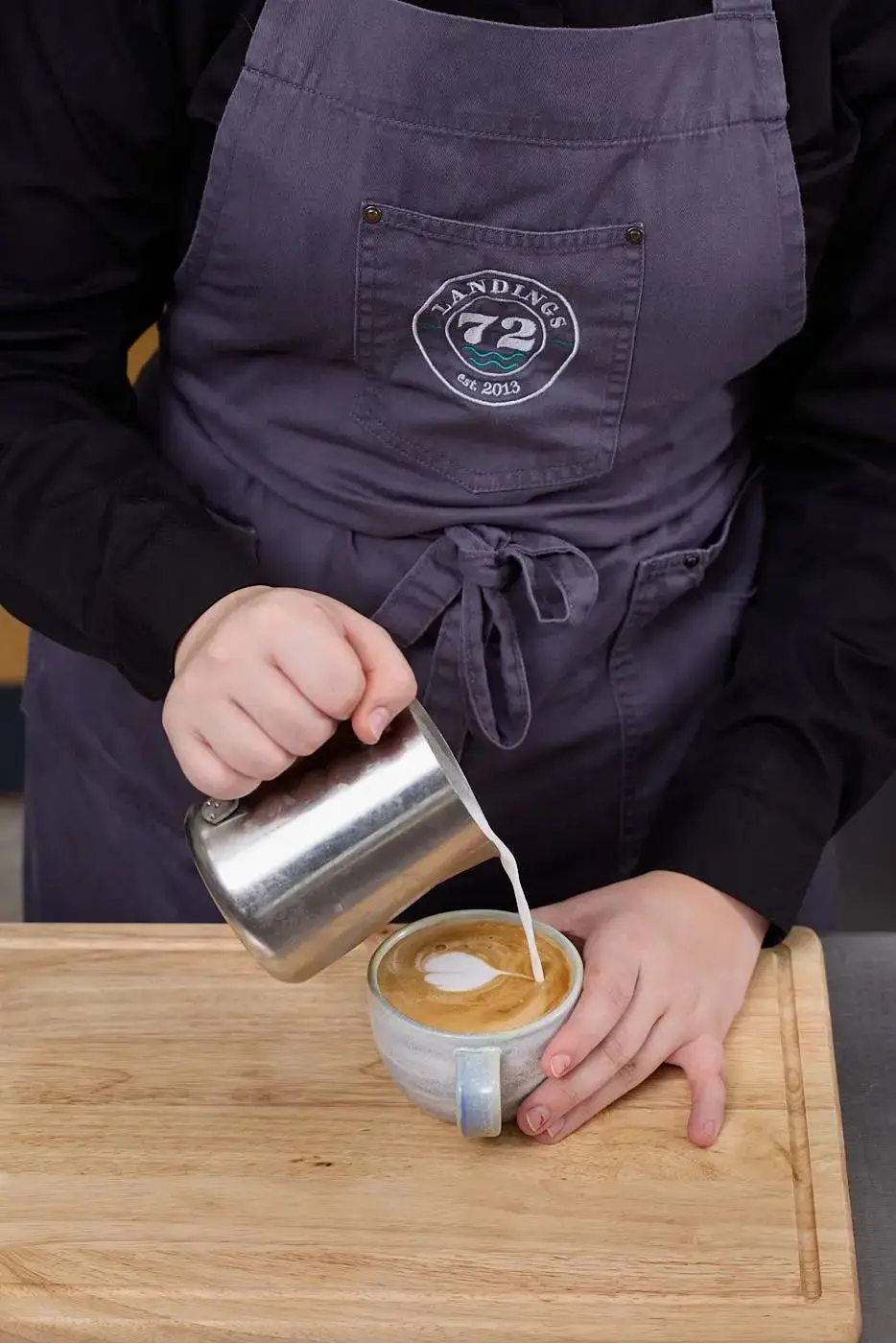 A person in a purple apron pouring milk into a cup of coffee on a wooden board.