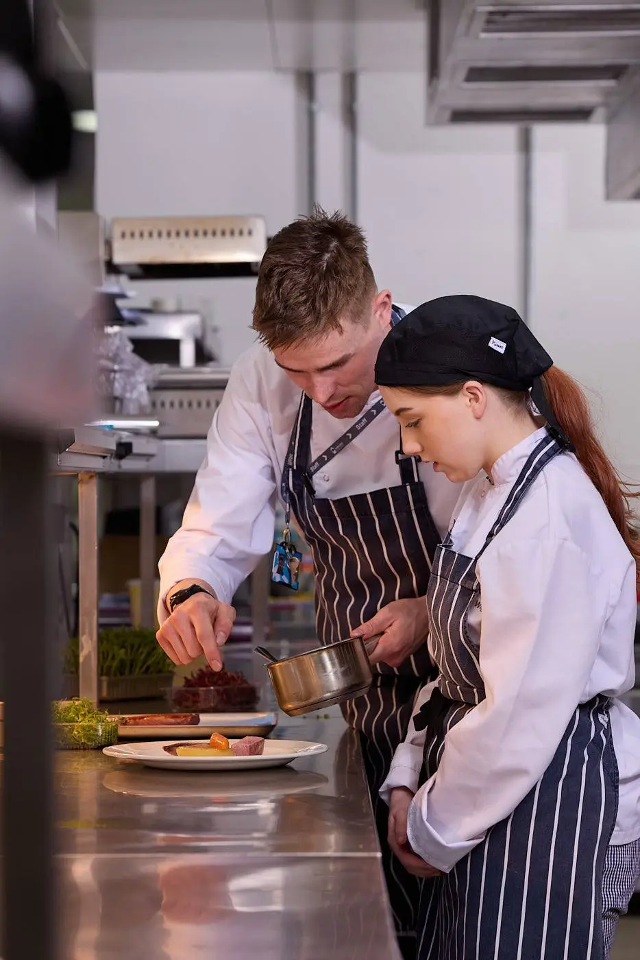 A chef and student in a kitchen, plating a dish with sauce. They are focused on presentation, wearing aprons and chef hats. Educational setting, culinary training.