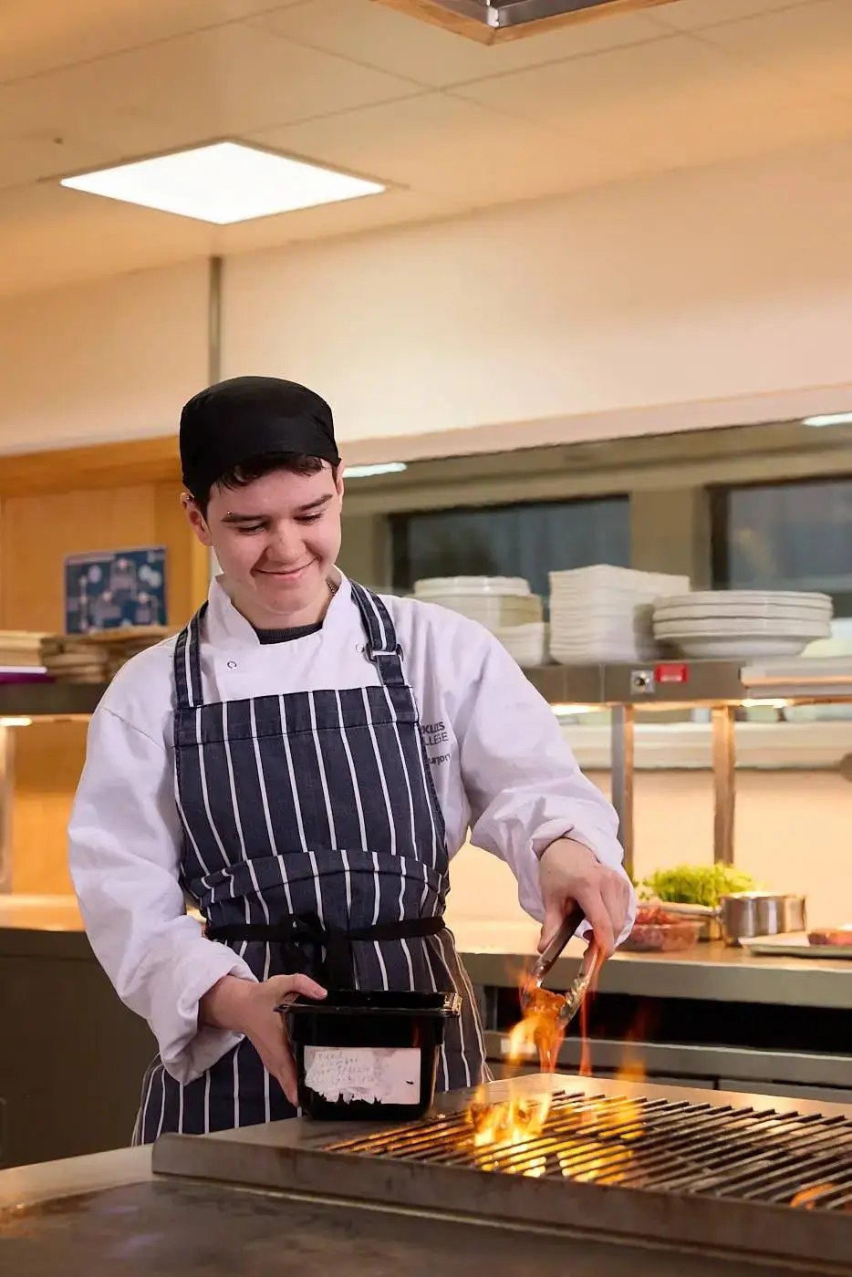 A student in a chef's uniform flambéing food on a grill in a college kitchen, smiling and focused on the culinary task.