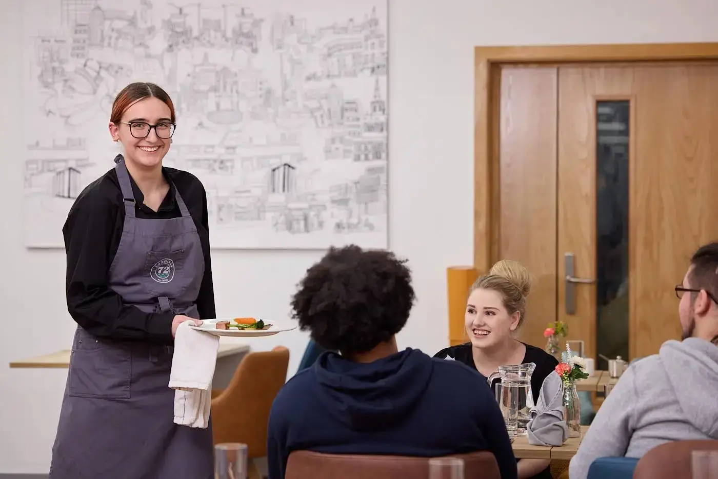 A student in an apron serves a dish to seated students in a college dining area with a cityscape mural.