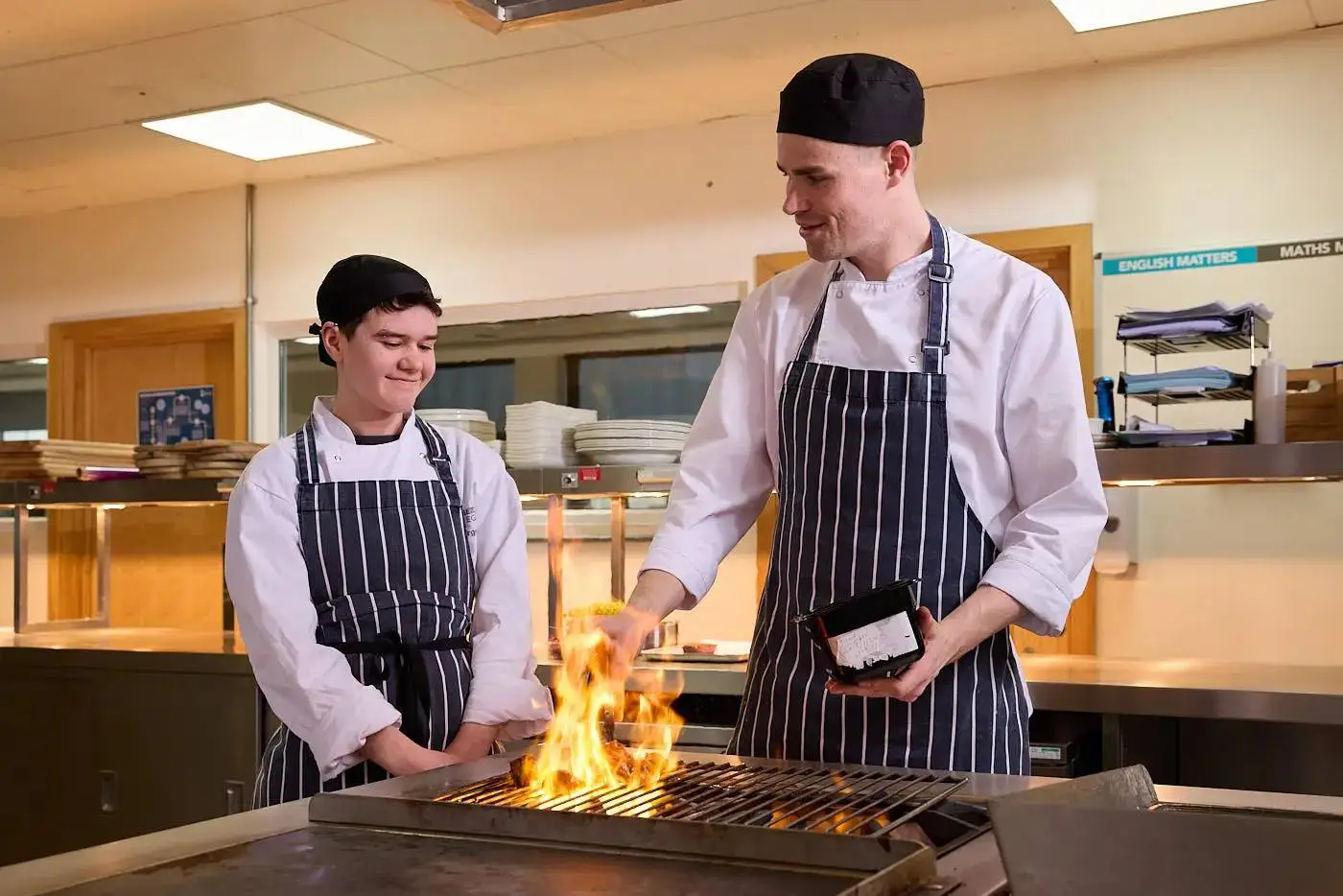 Two chefs in aprons and hats cooking on a grill in a college kitchen, with flames visible and plates stacked in the background.