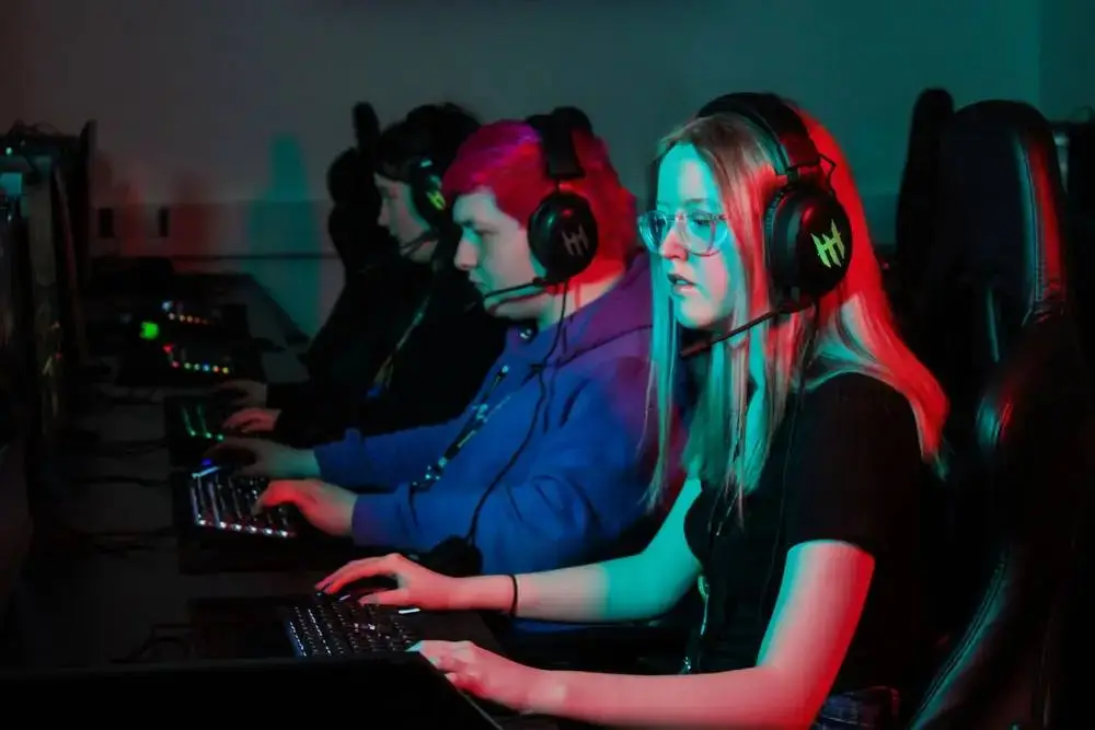 Students in gaming headsets at computers in a dimly lit room, focused on their screens.