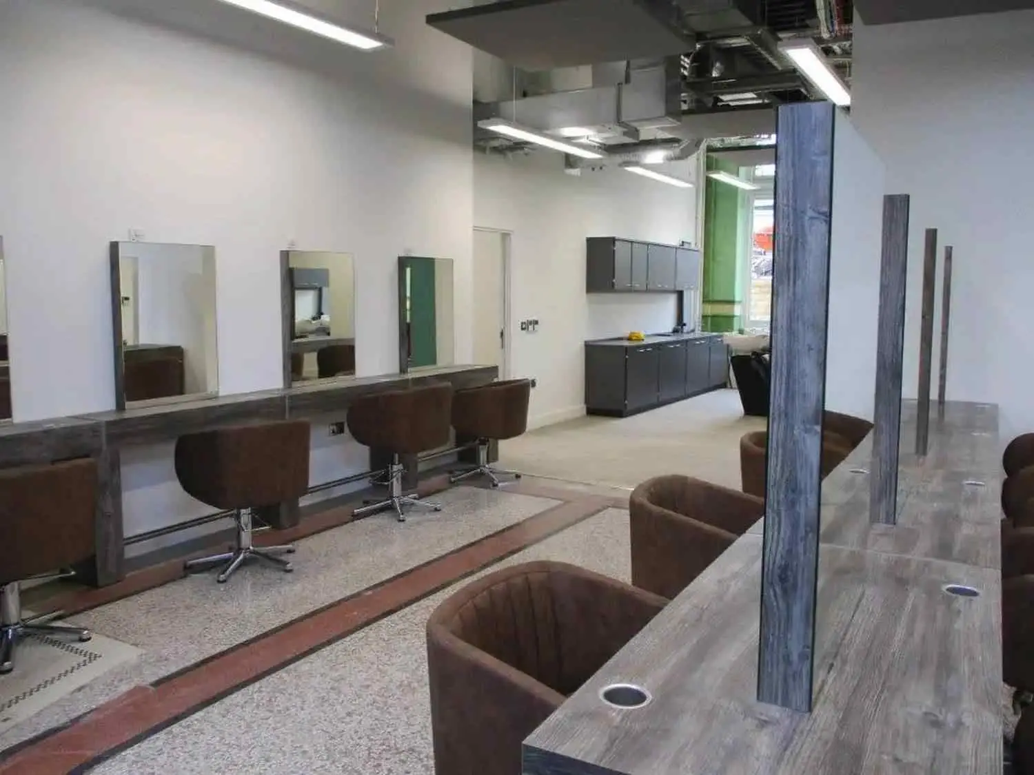 A modern college hair salon with brown chairs, mirrors, and grey cabinets, featuring exposed ductwork and ample lighting.