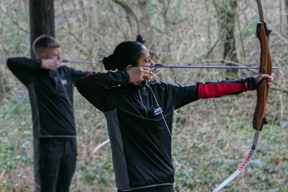 Two individuals practising archery outdoors, focusing on form and technique. They are wearing black sportswear, with one using a recurve bow. The setting is a natural, wooded area.