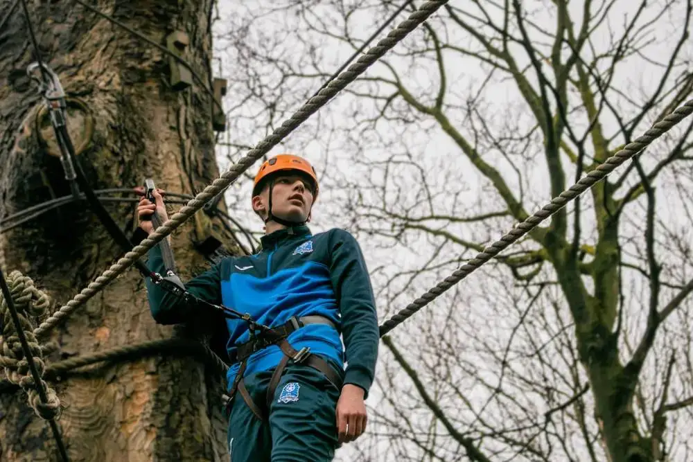 A person in a harness and helmet navigates a rope course in a tree, showcasing outdoor adventure and skill development in a natural setting.