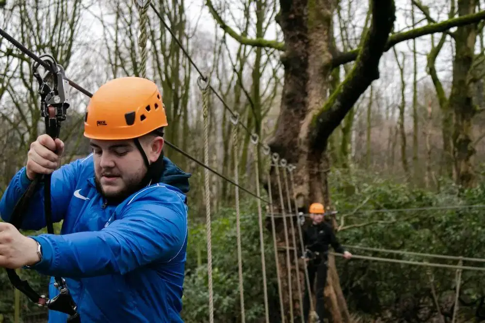 Two people in orange helmets and harnesses navigating a rope bridge in a wooded area, showcasing outdoor adventure and teamwork skills.