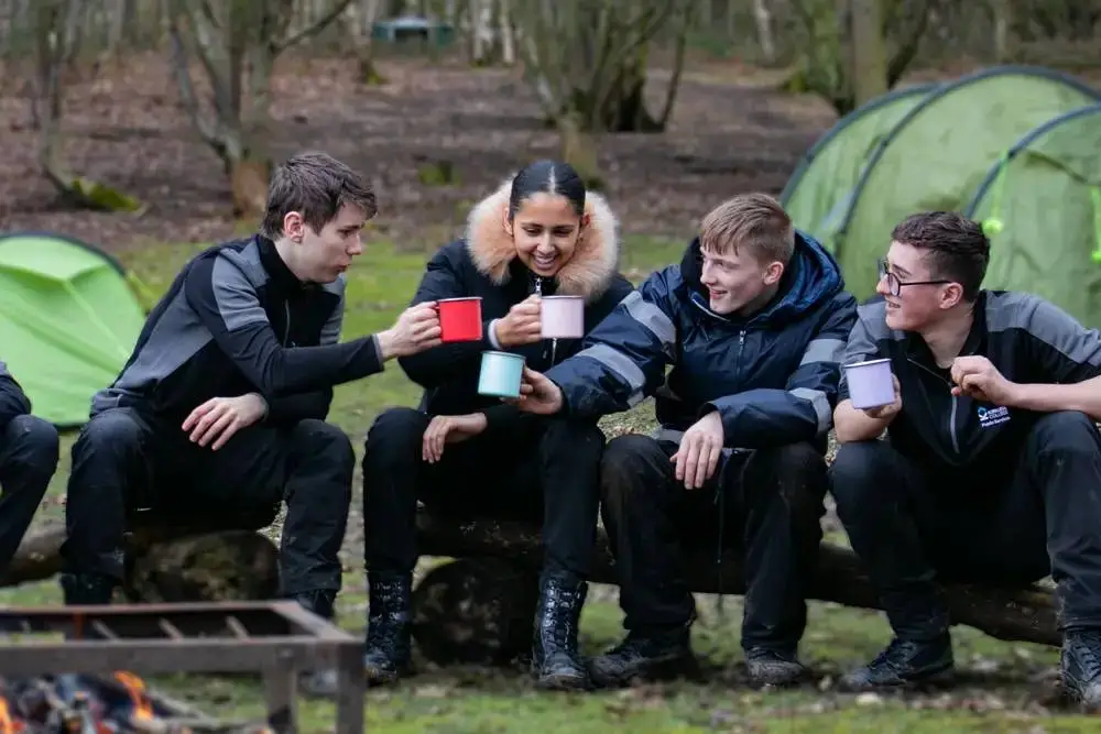Students in outdoor gear sitting around a campfire, holding colourful mugs, engaged in a group activity during a camping trip.
