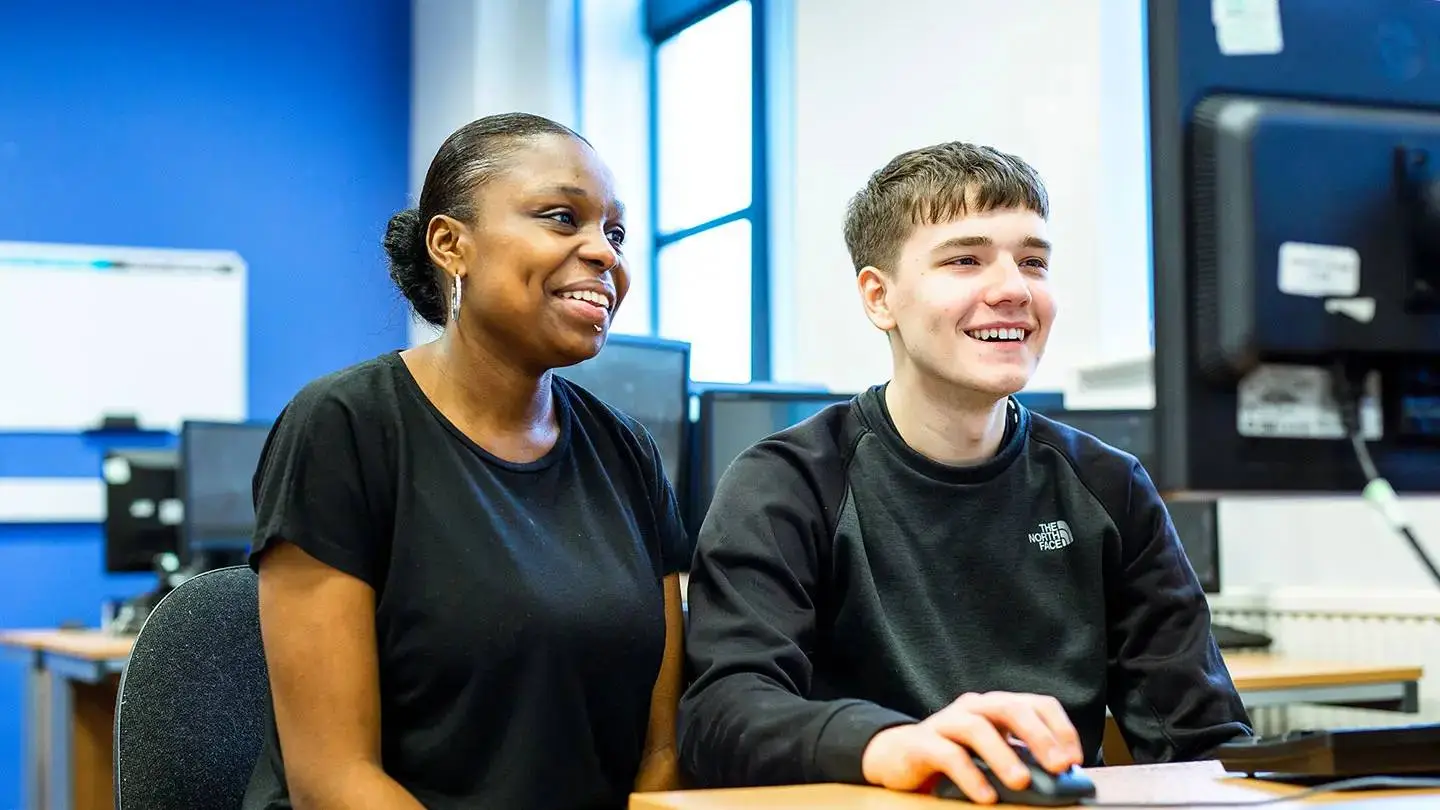 Two students smiling at a computer in a college classroom with blue walls and windows.