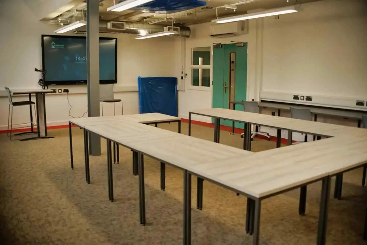 A college classroom with U-shaped tables, a large screen, and a projector. The room has a carpeted floor and fluorescent lighting.