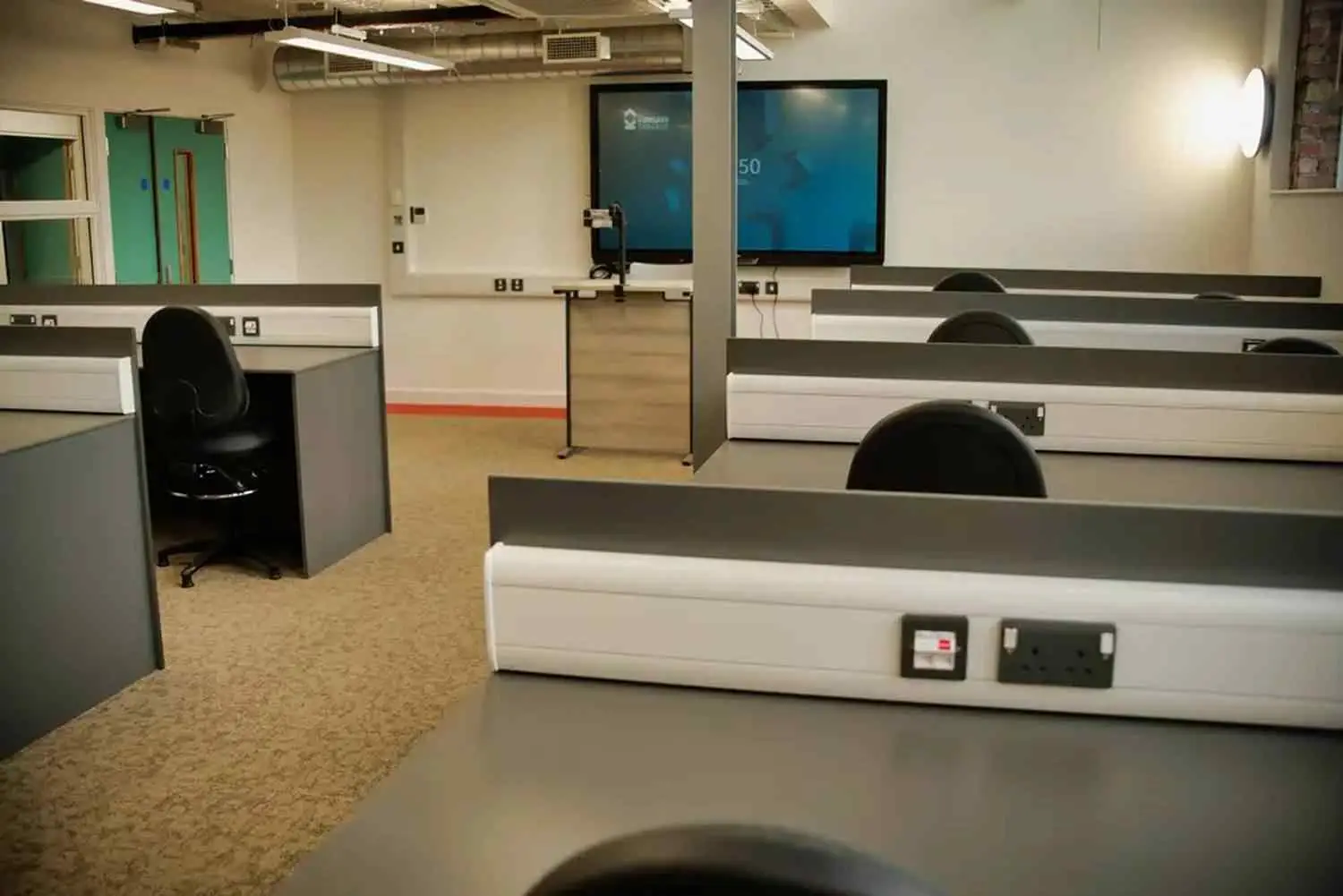 A college computer lab with rows of desks, black chairs, and large screens at the front. Power sockets and a carpeted floor are visible.