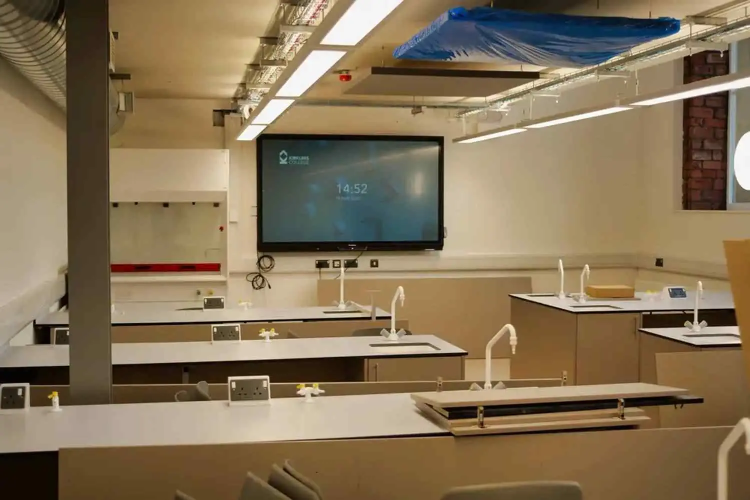 A university chemistry lab with white benches, sinks, and a large screen displaying the time. Blue safety cover above.