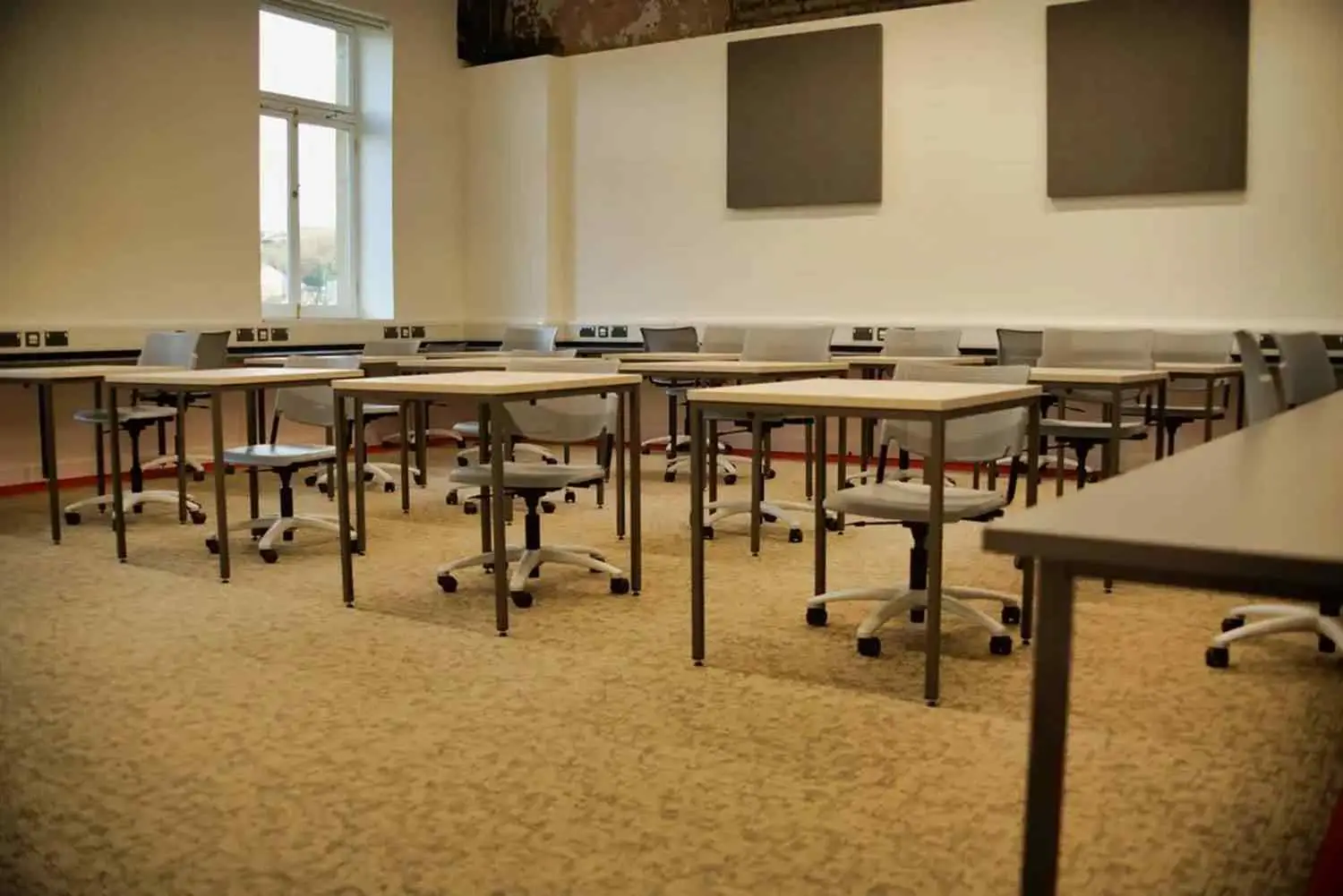 A university classroom with rows of desks and chairs, acoustic panels on the walls, and a window letting in natural light.