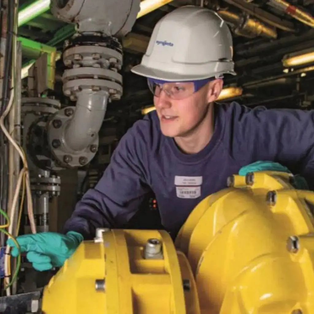 A person in a hard hat and gloves working on yellow machinery in an industrial setting.