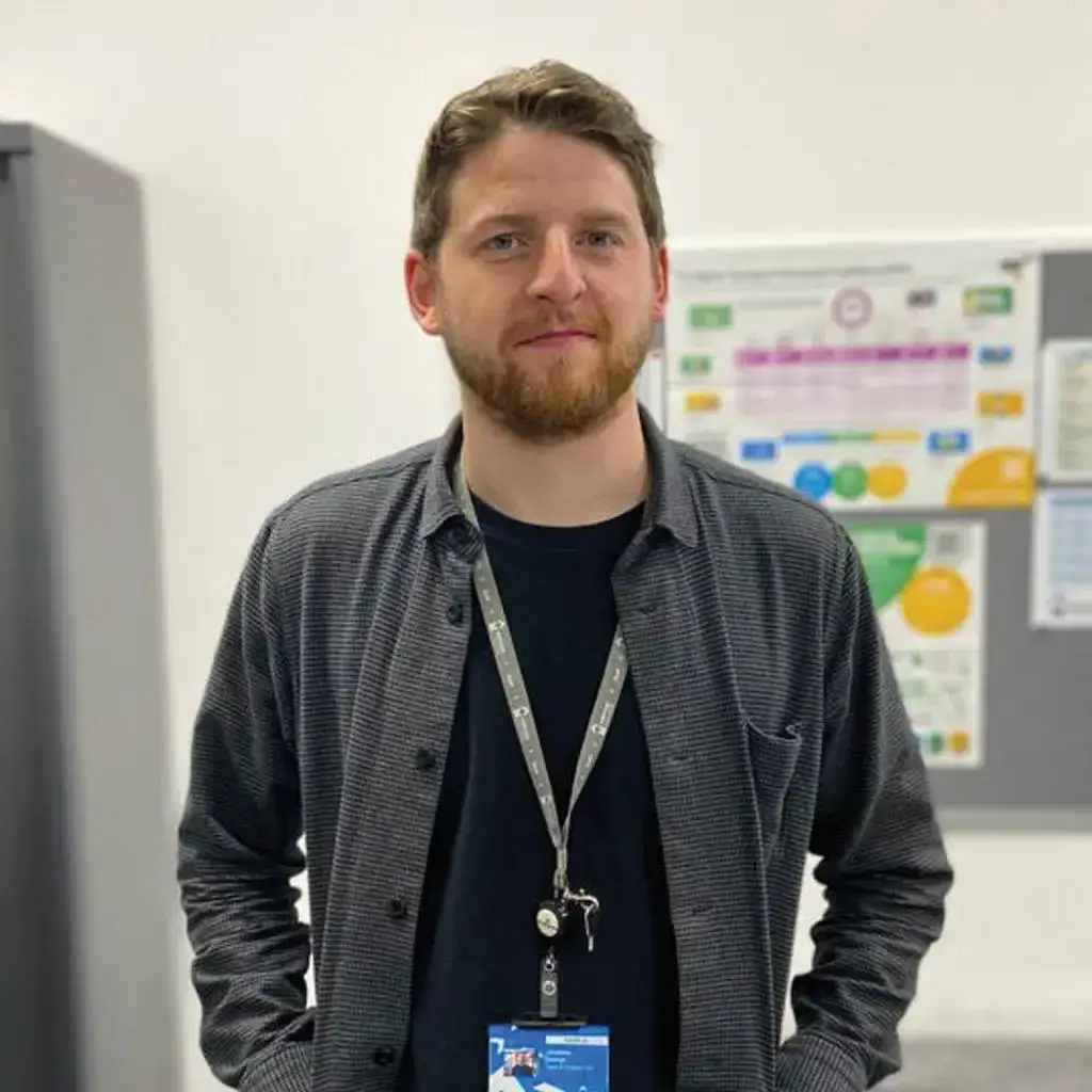 A man in a grey shirt with a lanyard stands in front of a whiteboard with colourful charts in a college classroom.