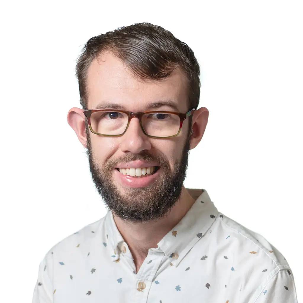 man with glasses and a beard smiling in a white shirt with small leaf patterns.