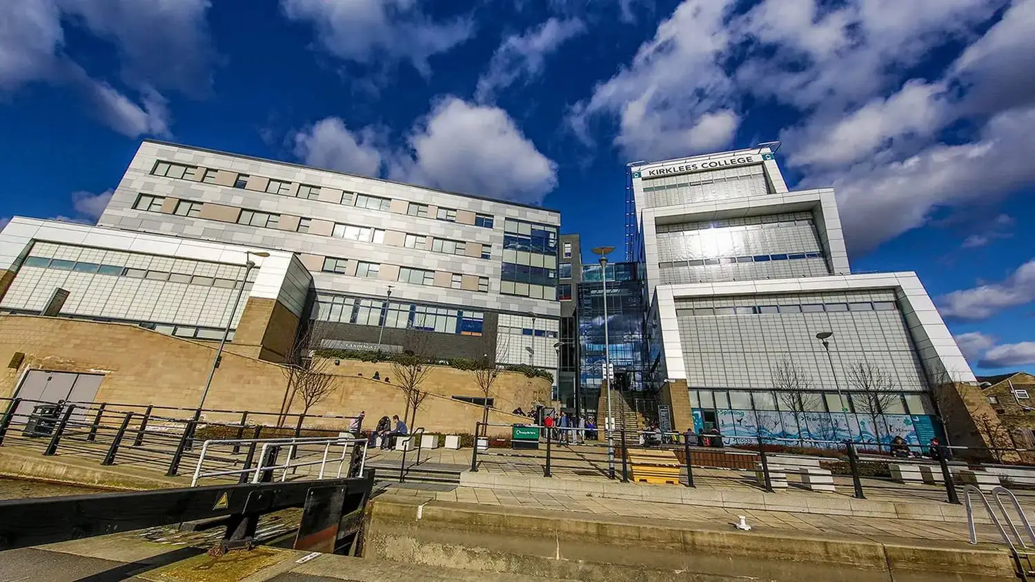 Modern college building with glass and brick facade under a blue sky with clouds. Students walking outside.