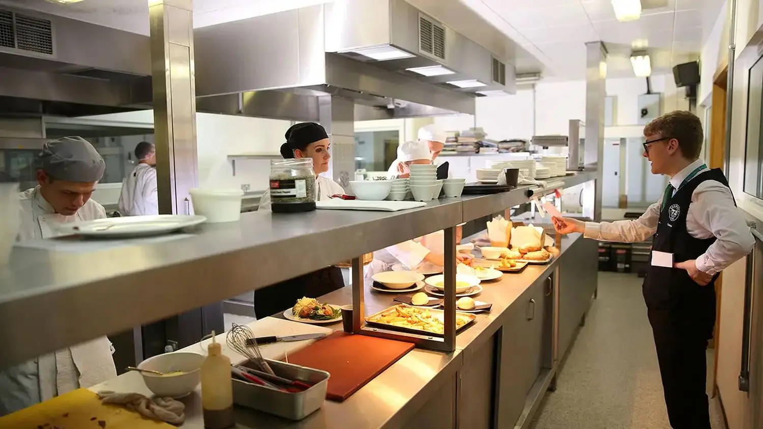 Culinary students in white uniforms serve dishes at a pass in a college kitchen, showcasing practical food preparation skills.