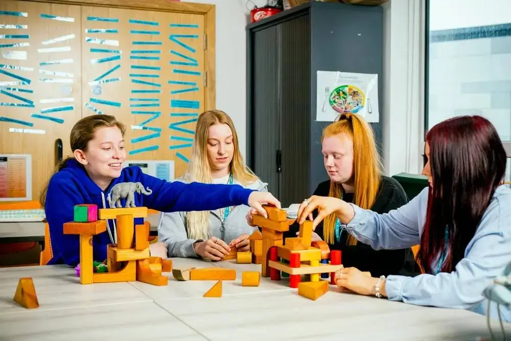 Four students collaboratively building a wooden structure on a table in a classroom setting.