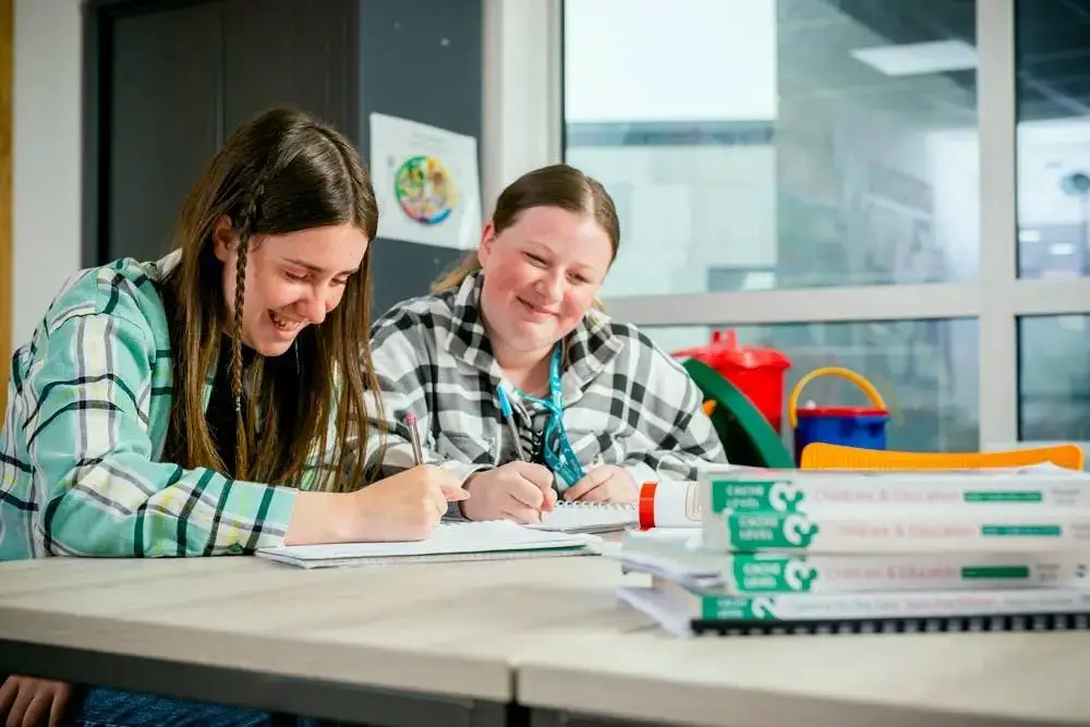 Two students in plaid shirts working together at a desk with notebooks and recycling bins in a bright classroom.