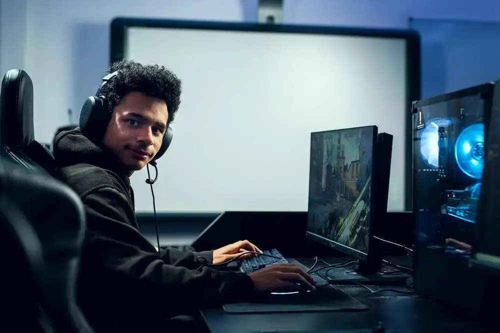 A student in a black hoodie wearing headphones sits at a desk, focused on a computer screen displaying a video game. A second monitor and a gaming PC are visible.
