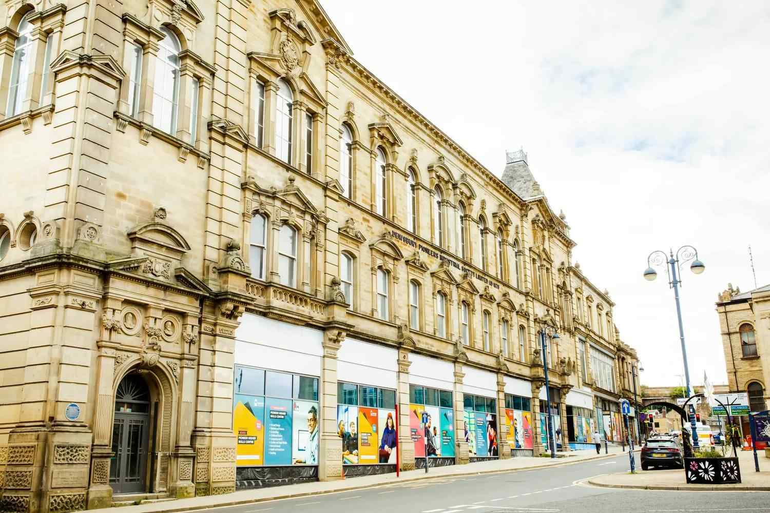 Historic stone building with arched windows and ornate details, housing a college. Modern posters on ground floor, street lamps, and a clear sky.