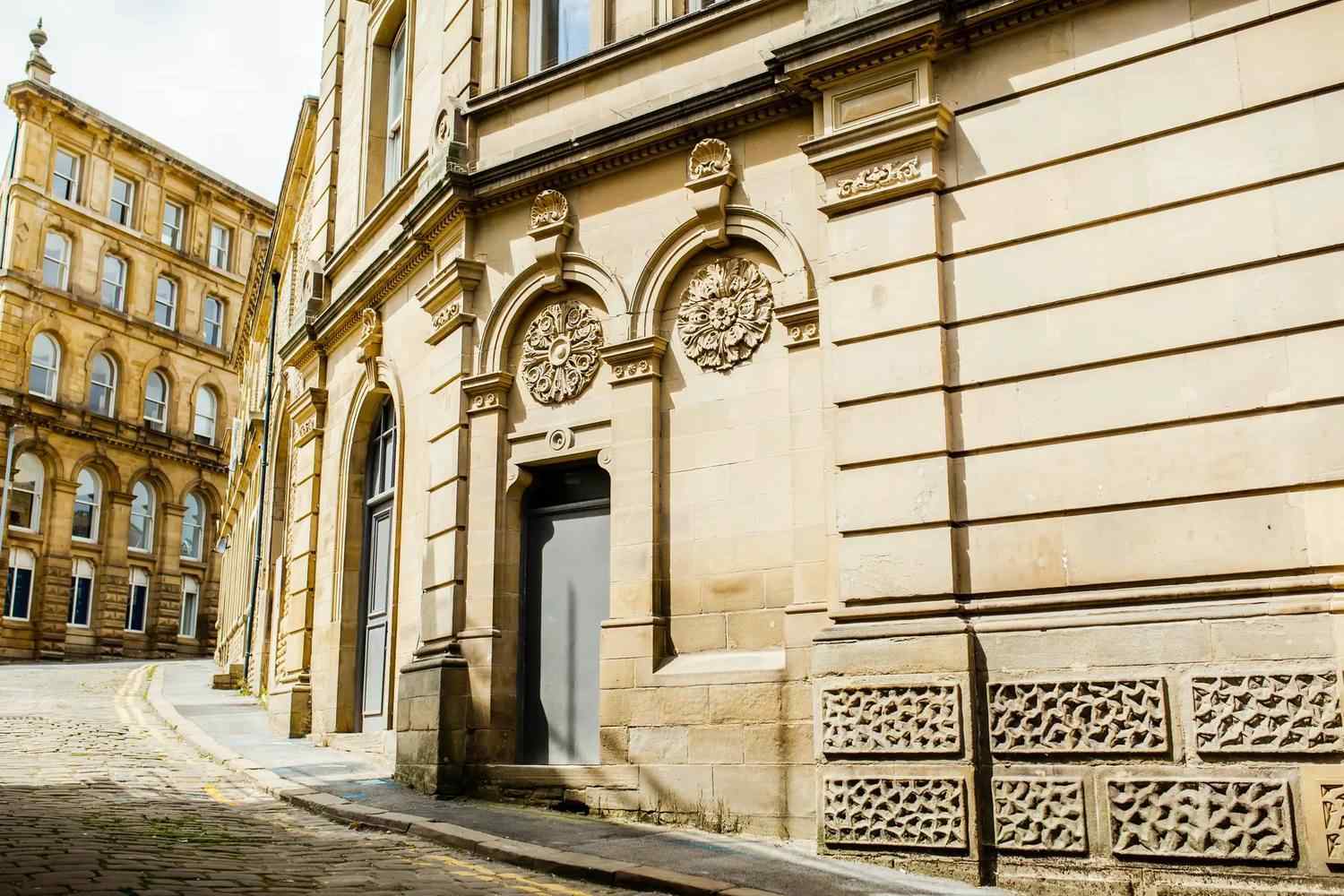 Historic stone building with arched windows and intricate carvings on a cobblestone street, reflecting classic British architecture.