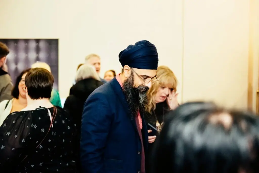 A man in a suit and turban stands in a room with other people, looking down at something in his hand.