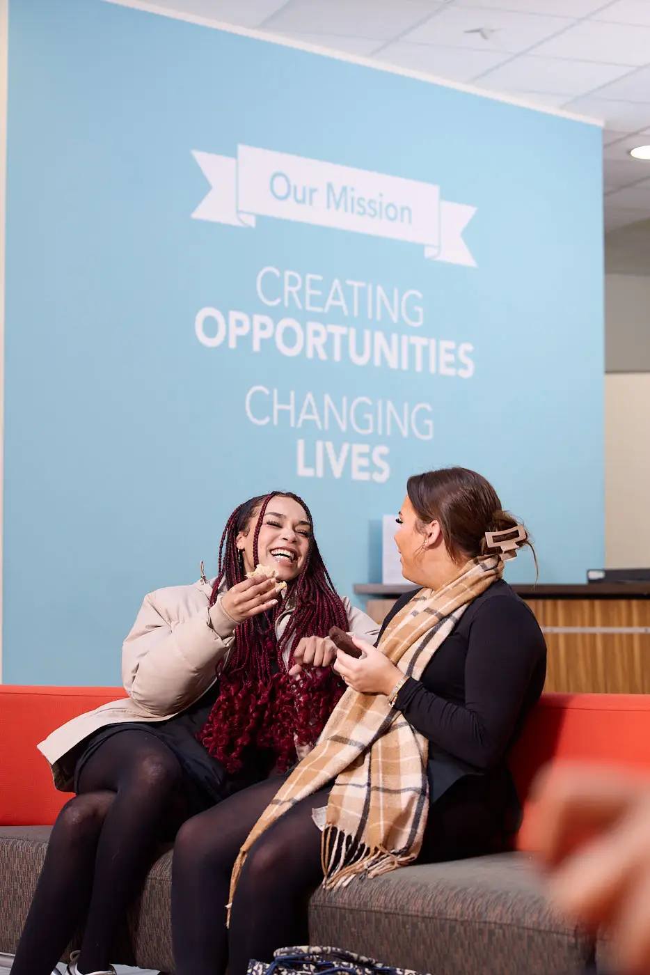 Two women sitting on a sofa, laughing and eating pastries. A blue wall behind them reads Our