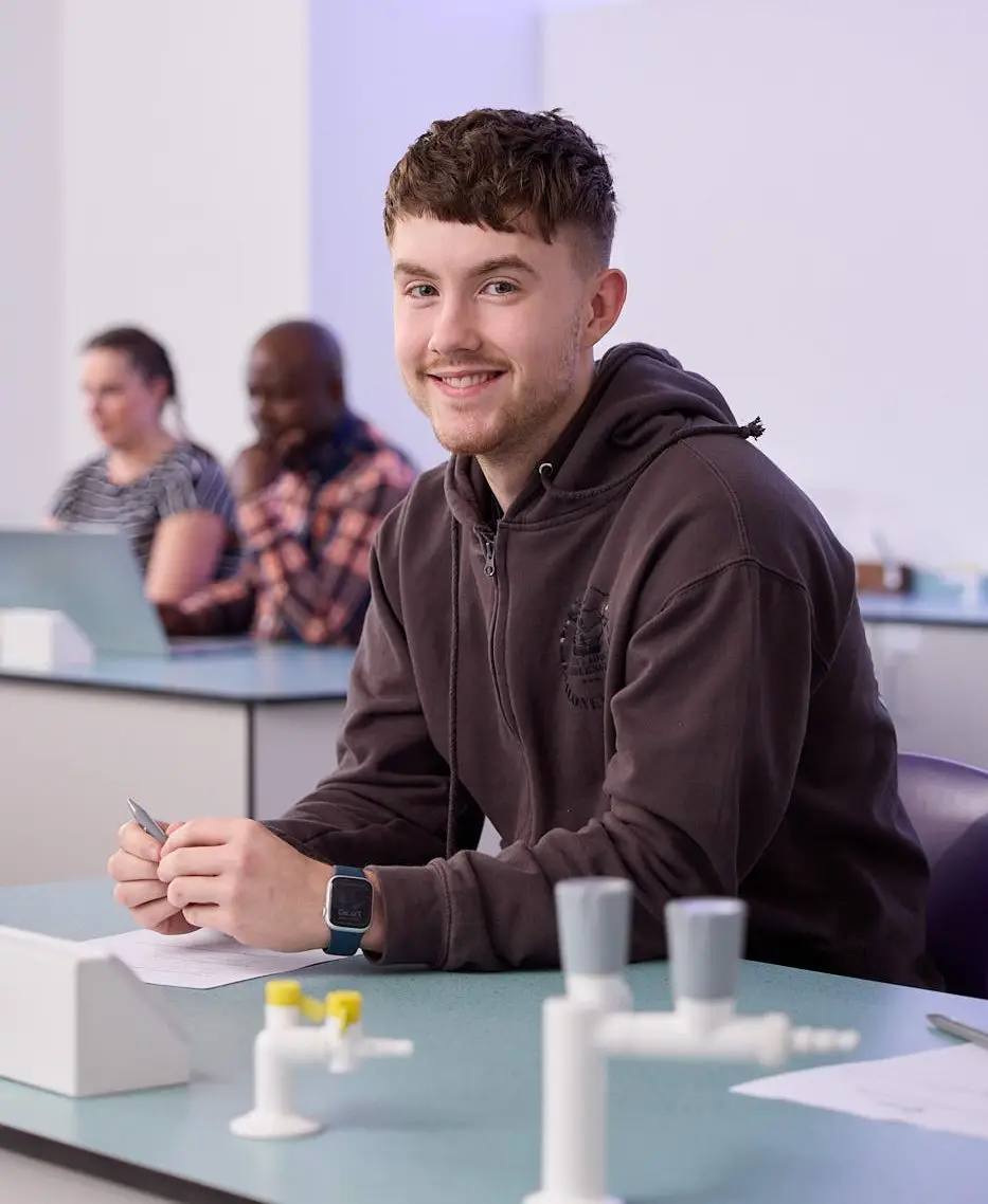 A student in a brown hoodie sits at a lab table, smiling and holding a pen, with lab equipment and other students in the background.