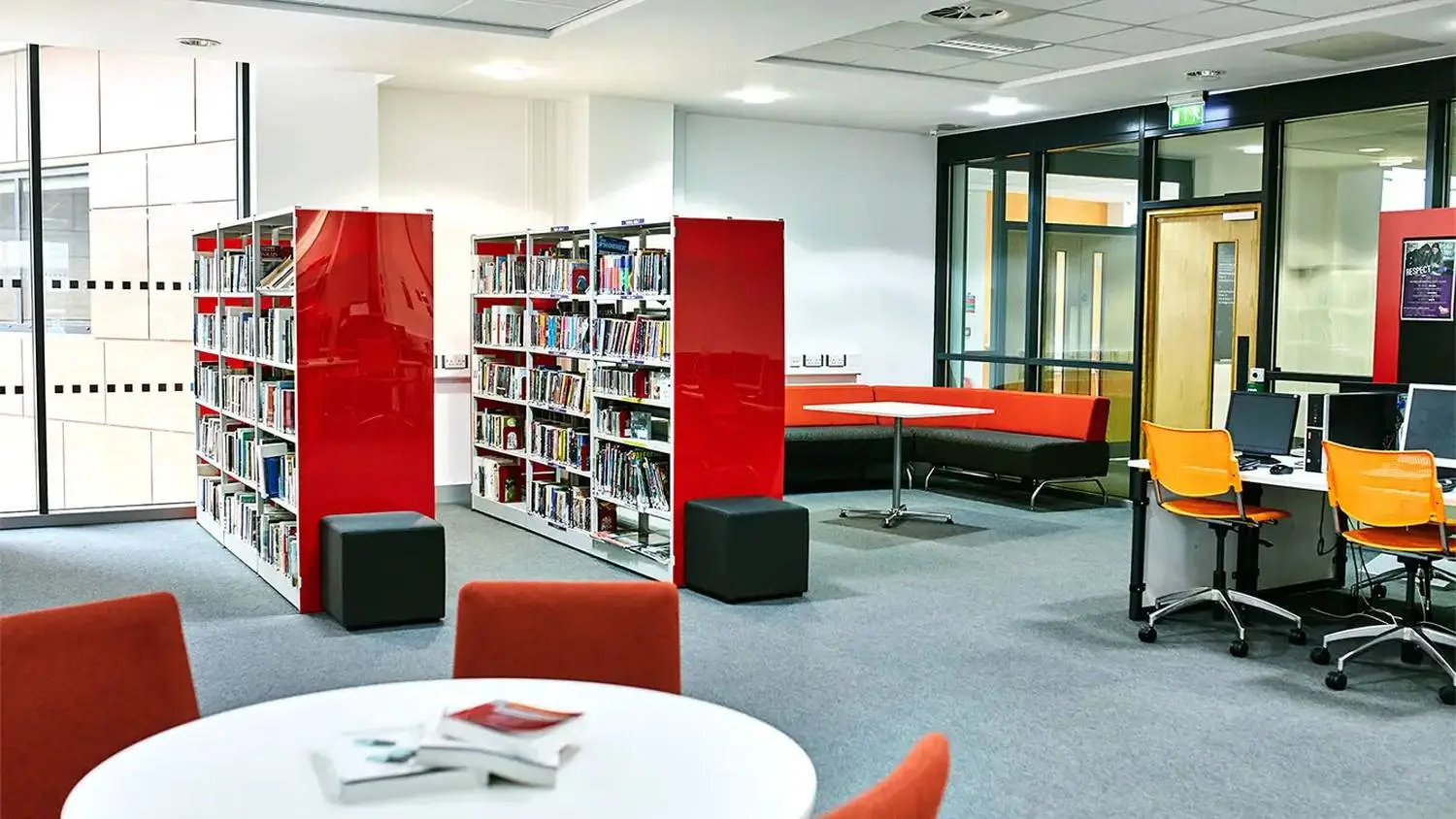 Modern college library with red bookshelves, orange seating, and computer workstations. Bright, open space with natural light.