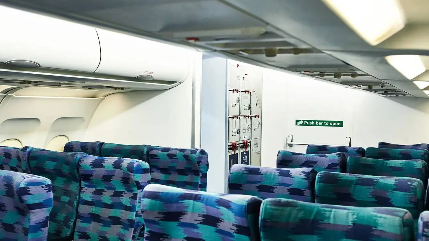 Train carriage interior with blue and green patterned seats, overhead luggage racks, and an exit sign. Push