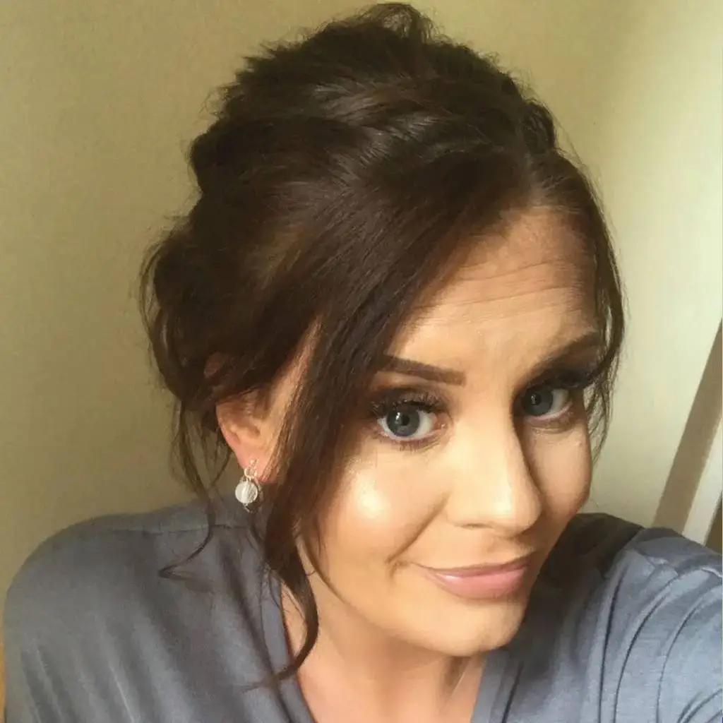 A woman with dark hair styled in an updo, wearing silver earrings, smiles and poses for a photo against a beige wall.