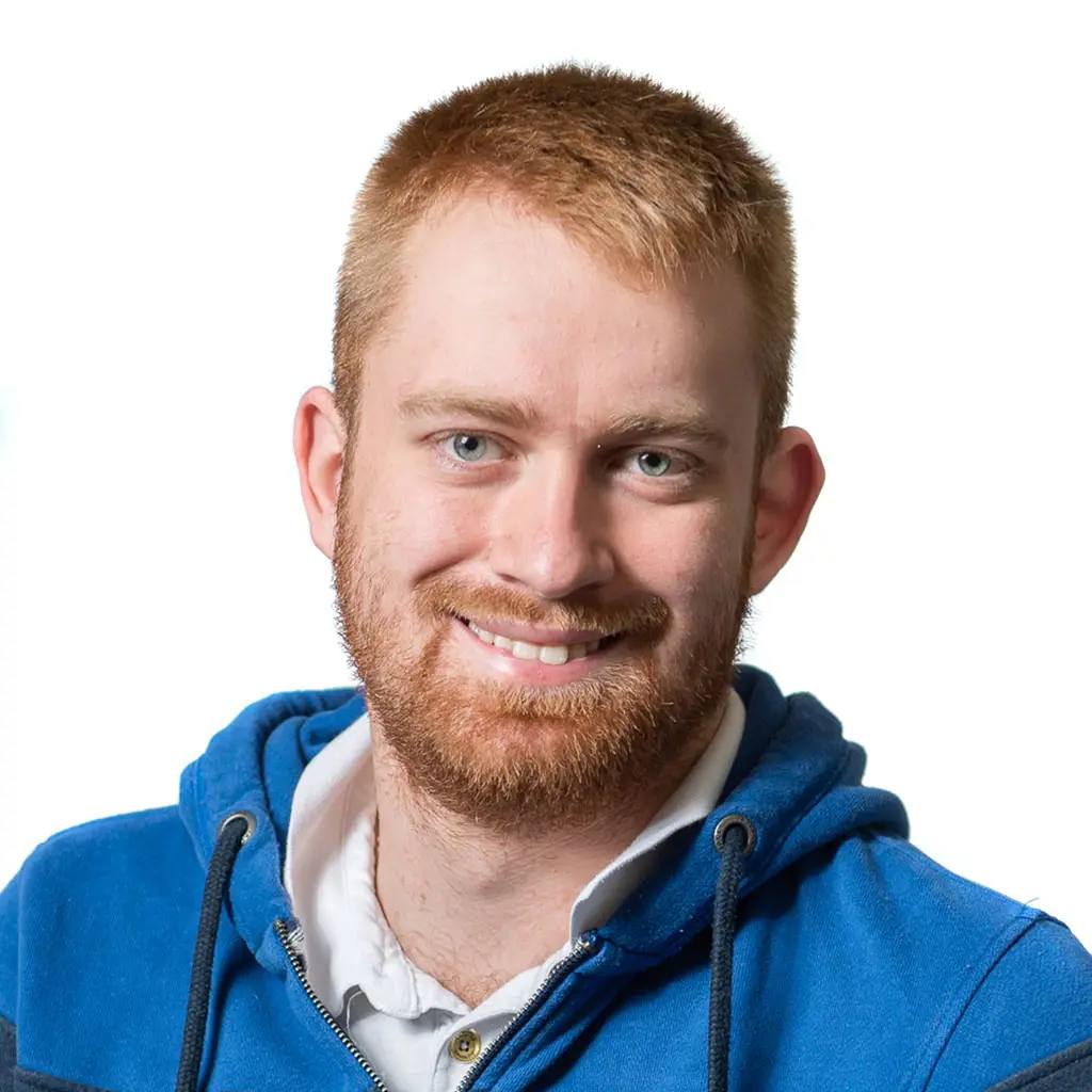 man with short red hair and a beard smiles at the camera, wearing a blue hoodie over a white shirt. He appears to be in a professional setting, possibly a college or university.