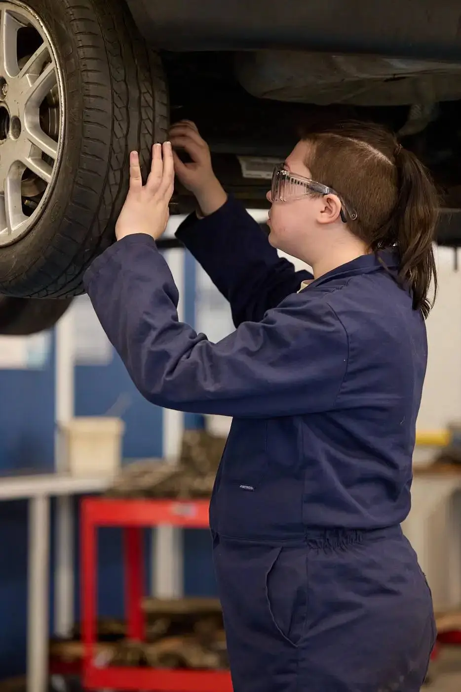A person in a blue jumpsuit and safety glasses working on a car wheel in a garage.