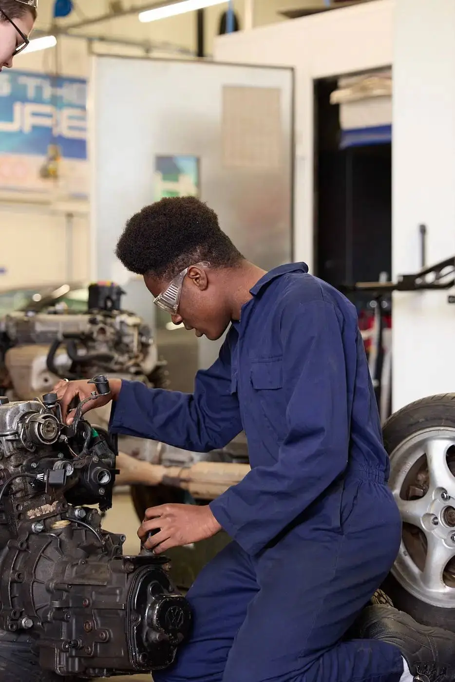 A person in a blue uniform and safety glasses working on a car engine in a workshop.