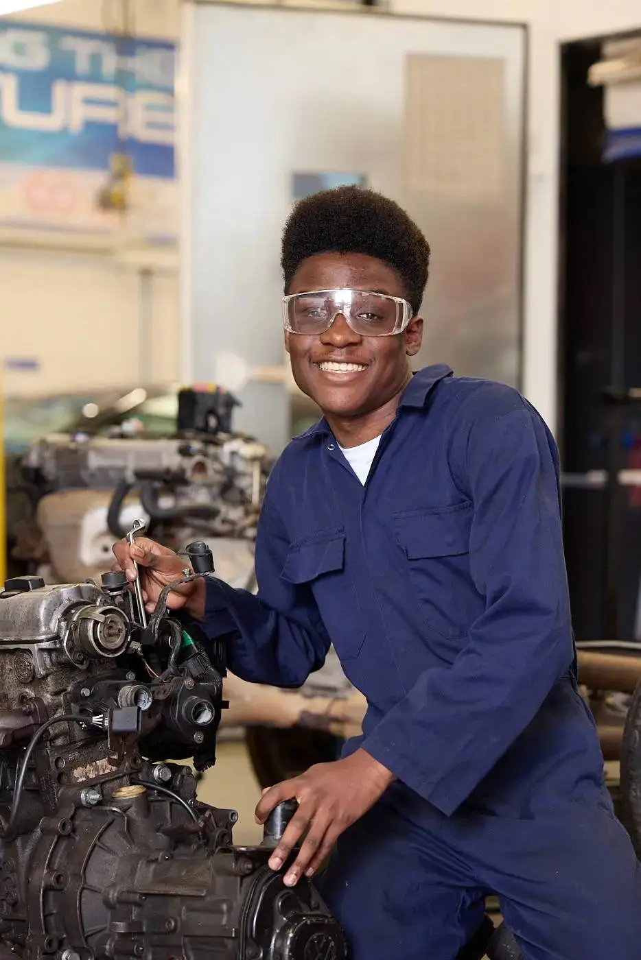 A person in blue overalls and safety glasses smiling and working on a car engine in a workshop.