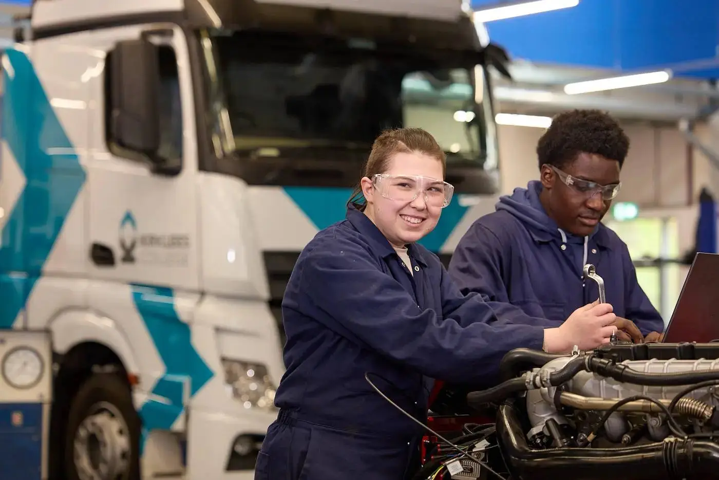 Two students in blue overalls and safety glasses working on a truck engine with a laptop in a college workshop.