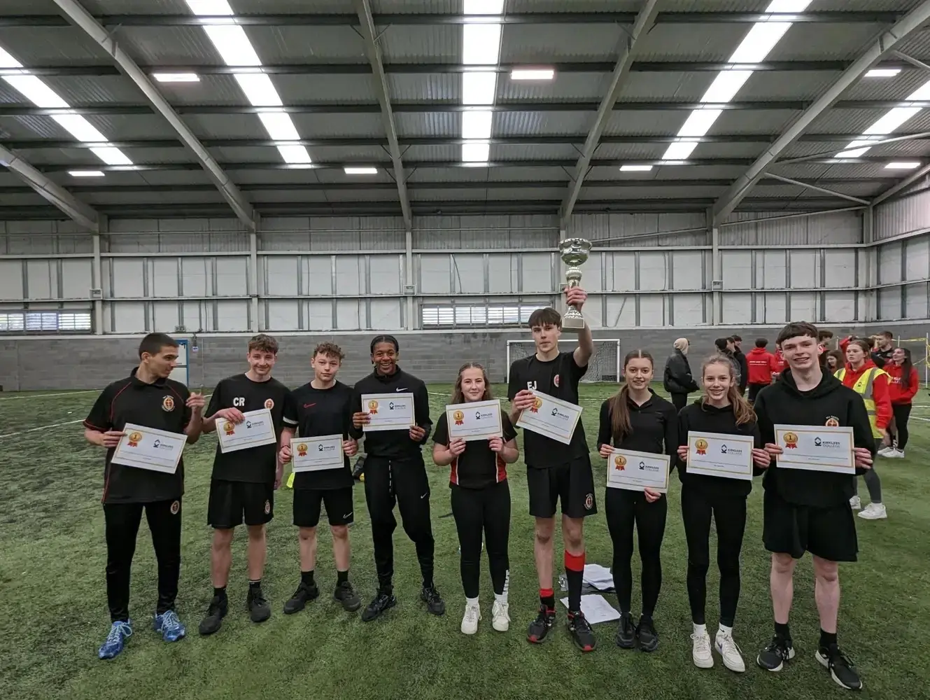 A group of college students in sports attire holding certificates and a trophy on an indoor football pitch, celebrating an achievement.