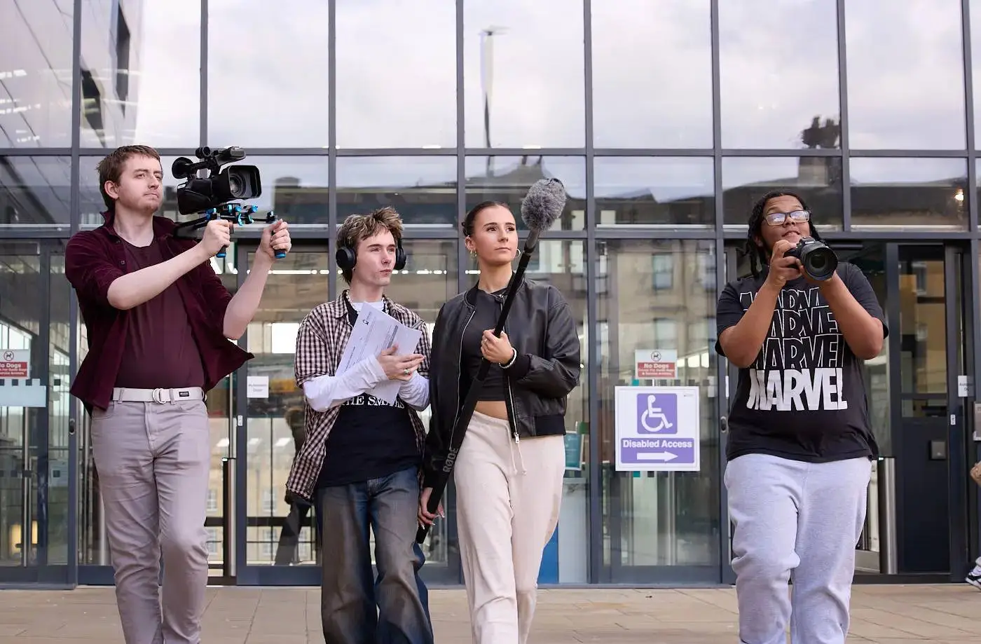 Four students filming outside a modern building with glass doors, holding camera and microphone equipment, engaged in a practical media project.