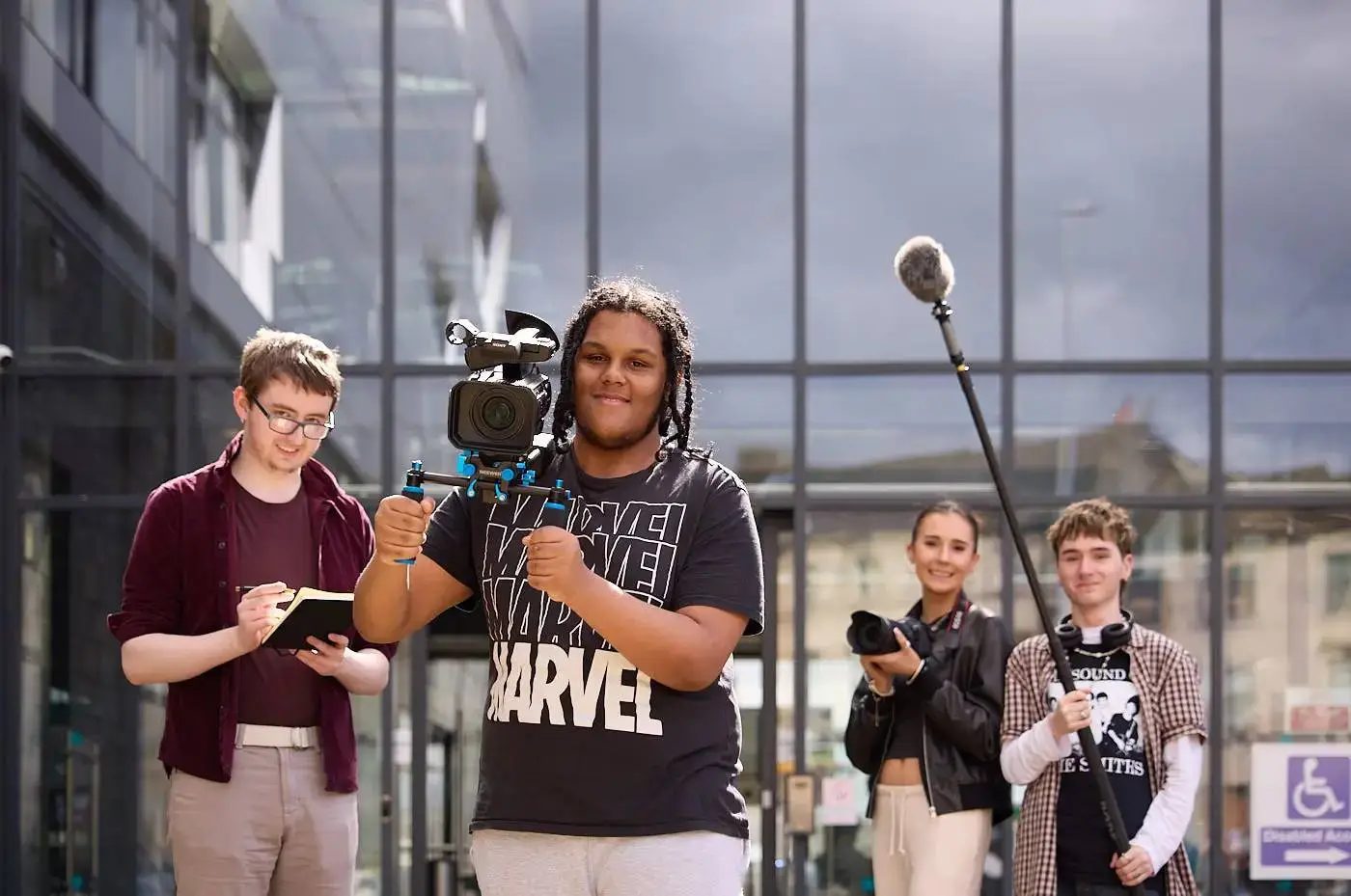 A group of students filming outside a modern building, holding cameras and microphones, engaged in a media project.