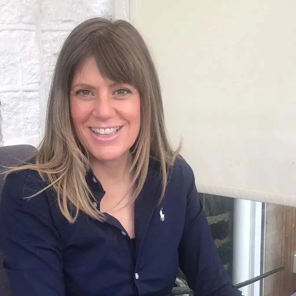 A woman in a navy shirt smiles at the camera, sitting in front of a white brick wall. She has long, straight hair with lighter highlights.