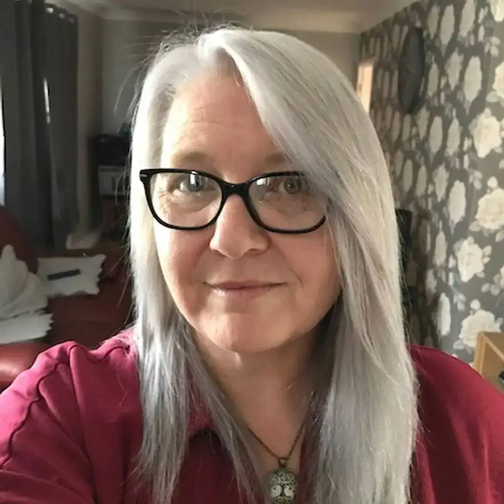 A woman with long white hair and glasses smiles at the camera, wearing a red blouse and a necklace with a circular pendant.