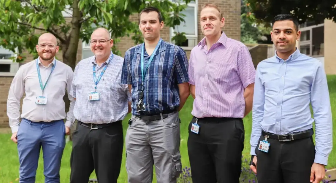 Five men in collared shirts and lanyards stand outdoors on a college campus, smiling and posing for a photo.