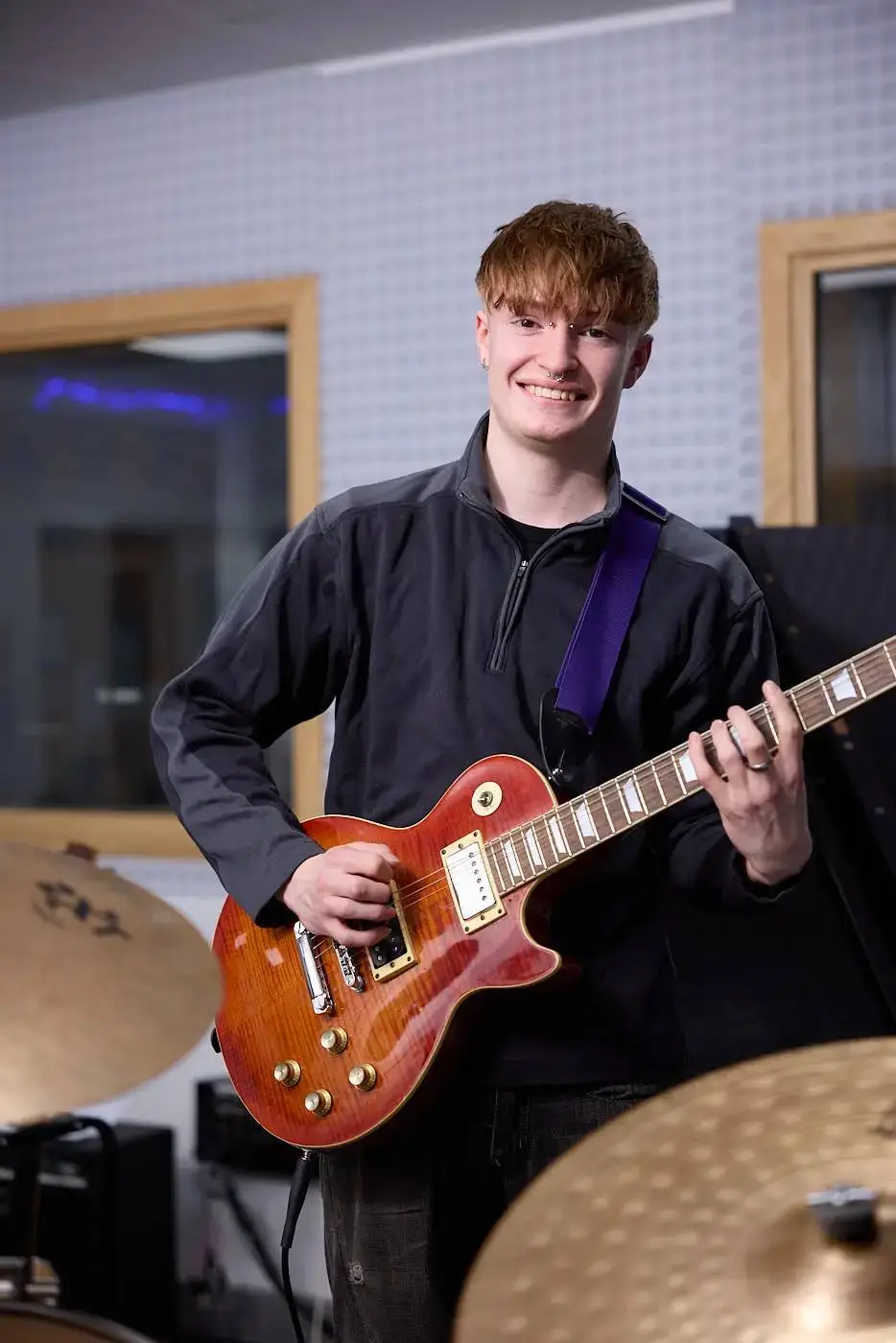 A student smiling and holding a red electric guitar in a music studio.