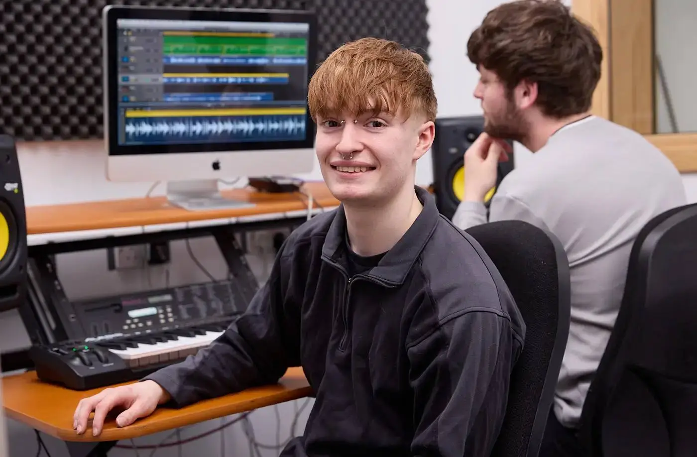 Two students in a sound recording studio, one smiling at the camera beside a computer and keyboard, the other focused on the screen.